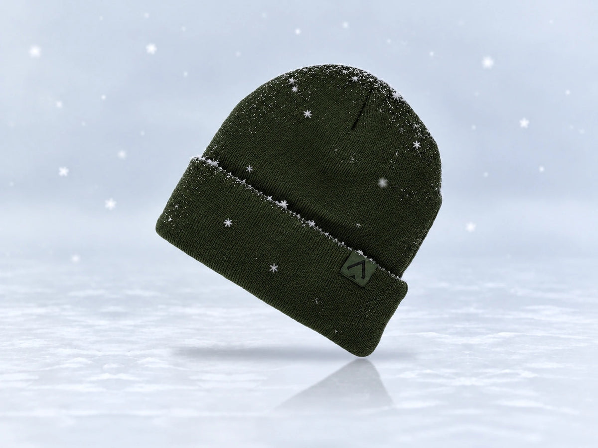 All In Lid Forest Green cuffed beanie in forest green knit with a black spade logo on a tonal green patch, angled view with a light dusting of snow and falling flakes against a pale gray background.