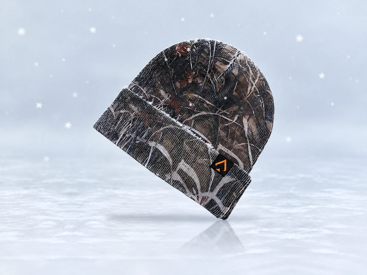 All In Lid Stealth Camo cuffed beanie in woodland camouflage knit with an orange spade logo on a black patch, angled view with a light dusting of snow and falling flakes against a pale gray background.