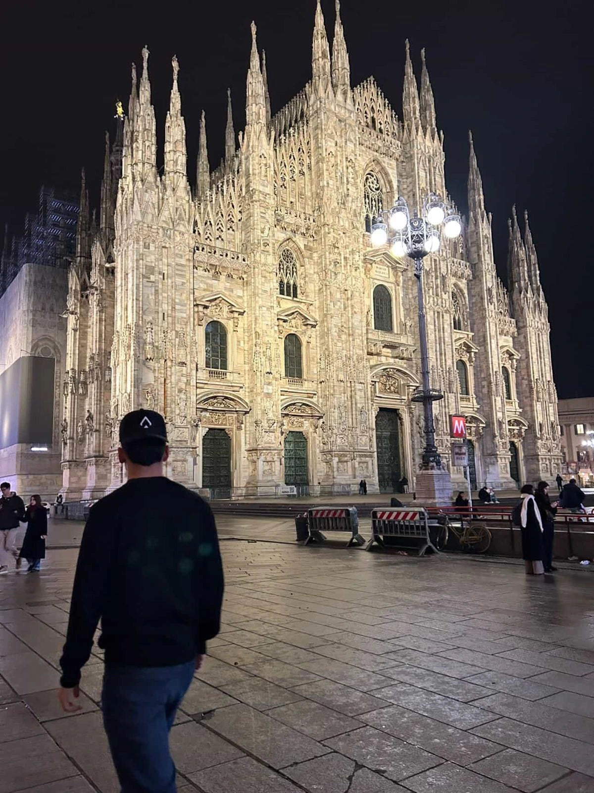 A man seen from behind wearing a black All In Lid lifestyle cap, walking toward a large, historic European building at night.