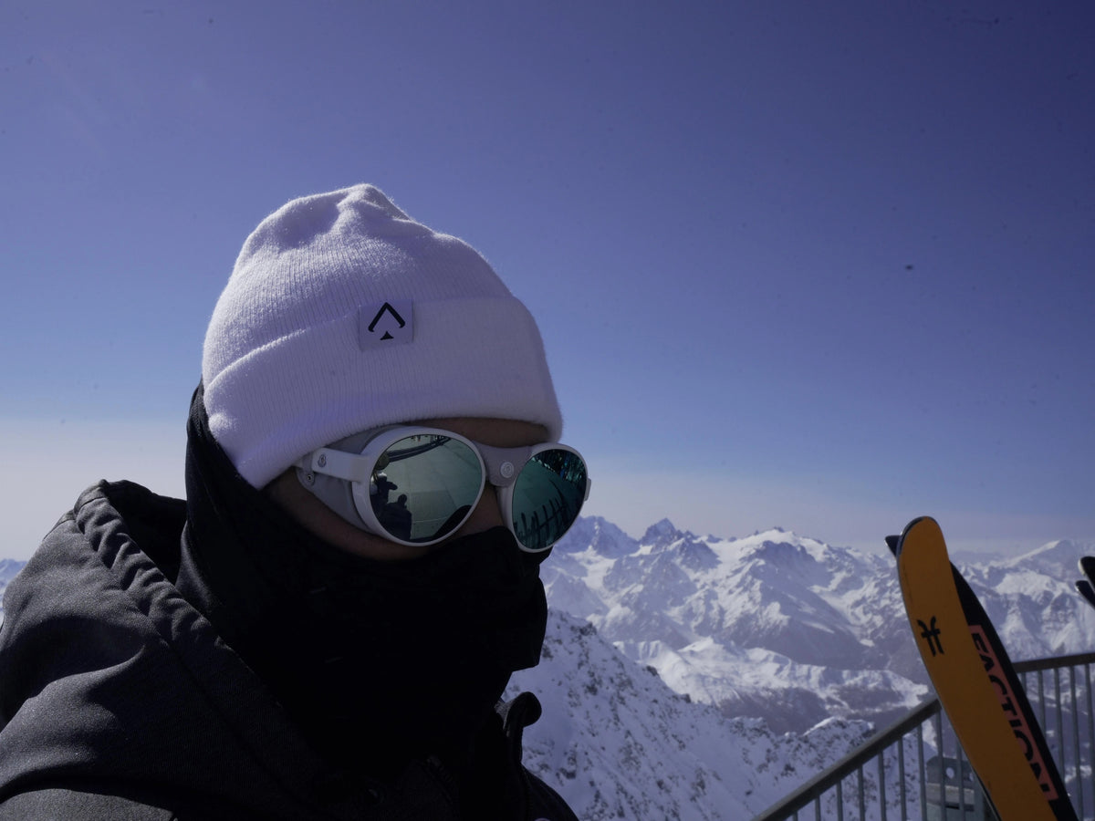 A close-up of a person in white All In Lid knit beanie and mirrored goggles with a view of a snowy mountain range.