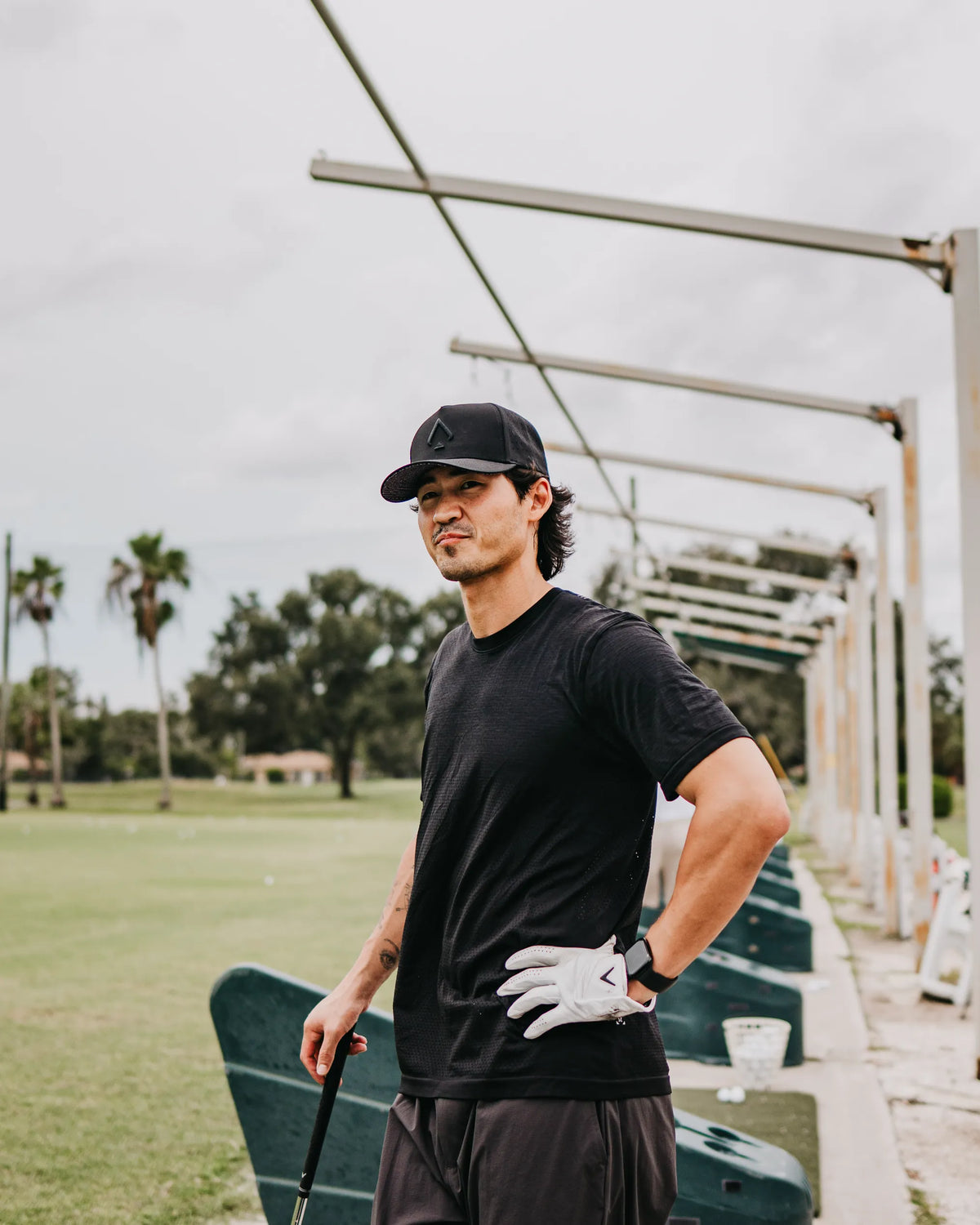 A man wearing a black t-shirt, black cap, and white golf glove stands on a golf driving range holding a club.
