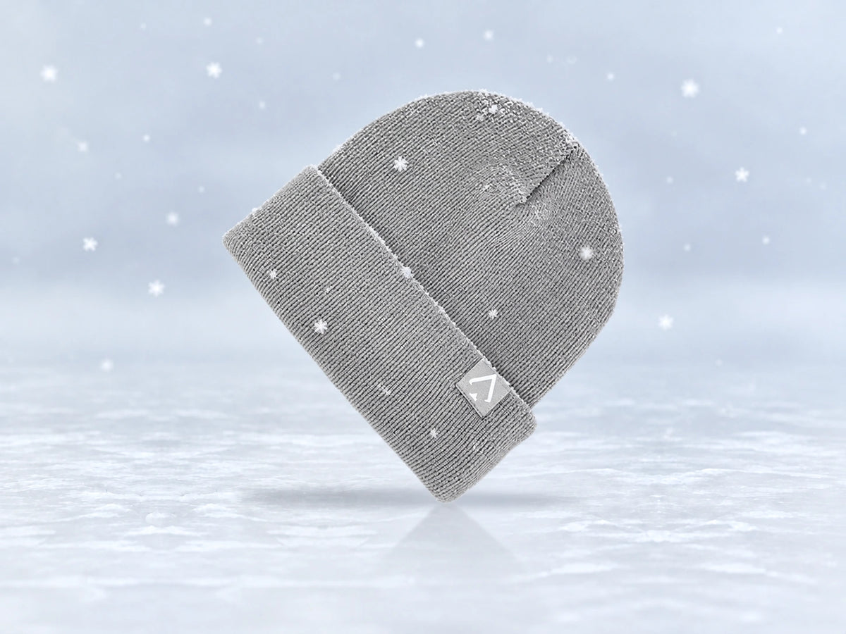 All In Lid Ash Gray cuffed beanie in light gray knit with a white spade logo on a light gray patch, angled view with a light dusting of snow and falling flakes against a pale gray background.