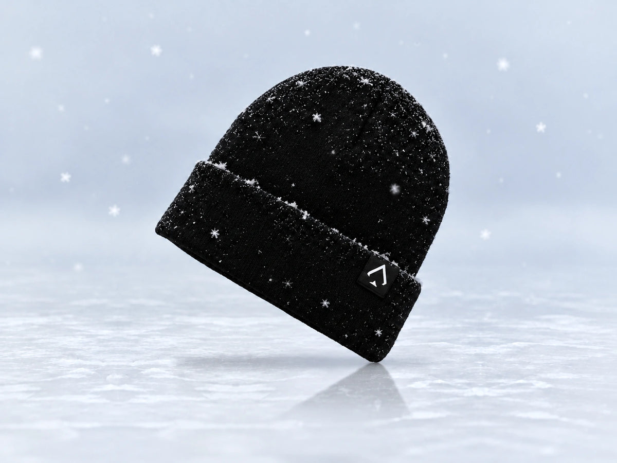 All In Lid Jet Black cuffed knit beanie in an angled view with a light dusting of snow, a white spade logo on a black patch, and scattered falling snowflakes against a pale gray background.