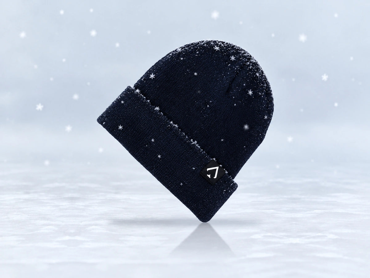 All In Lid Midnight Navy cuffed beanie in navy knit with a white spade logo on a black patch, angled view with a light dusting of snow and falling flakes against a pale gray background.