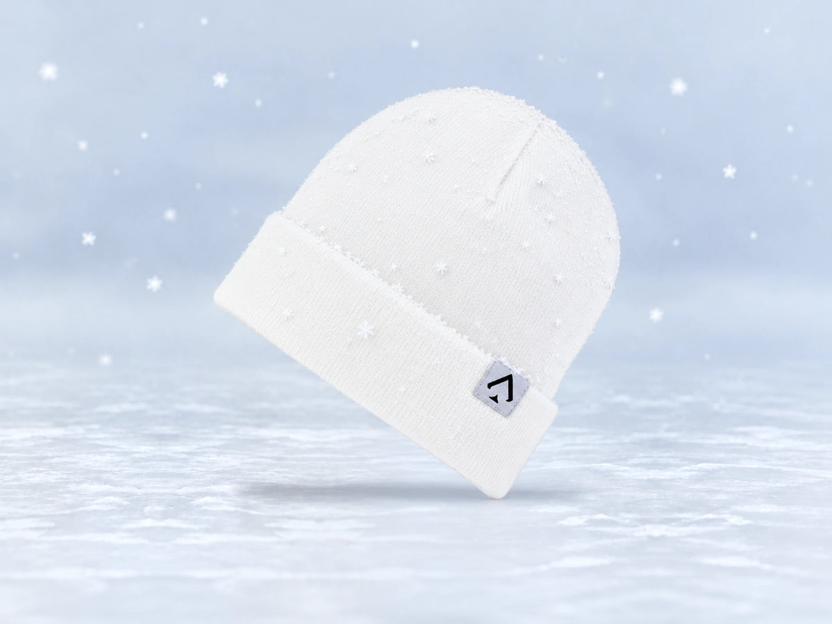 All In Lid Snow White cuffed beanie in white knit with a black spade logo on a light gray patch, angled view with a light dusting of snow and falling flakes against a pale gray background.