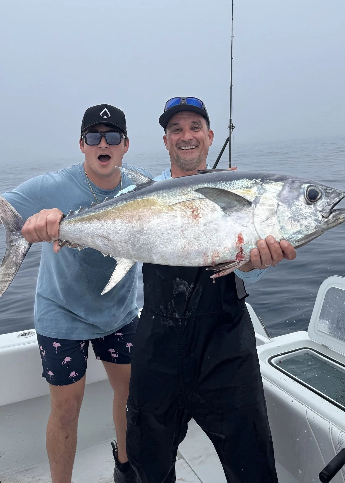 Two men on a fishing boat holding a large tuna fish, with one man wearing a black All In Lid performance hat.